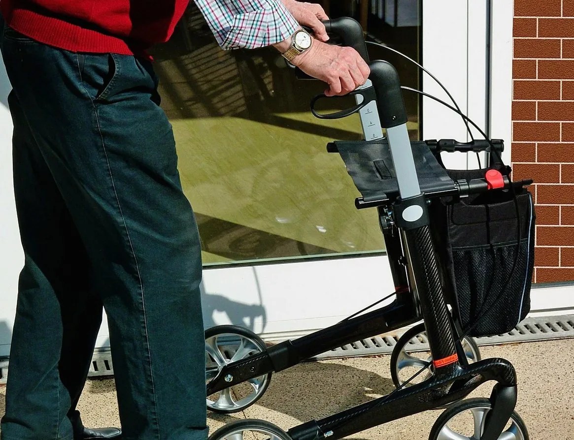 Person with a wheeled walking frame beside a glass door, shown from the waist down