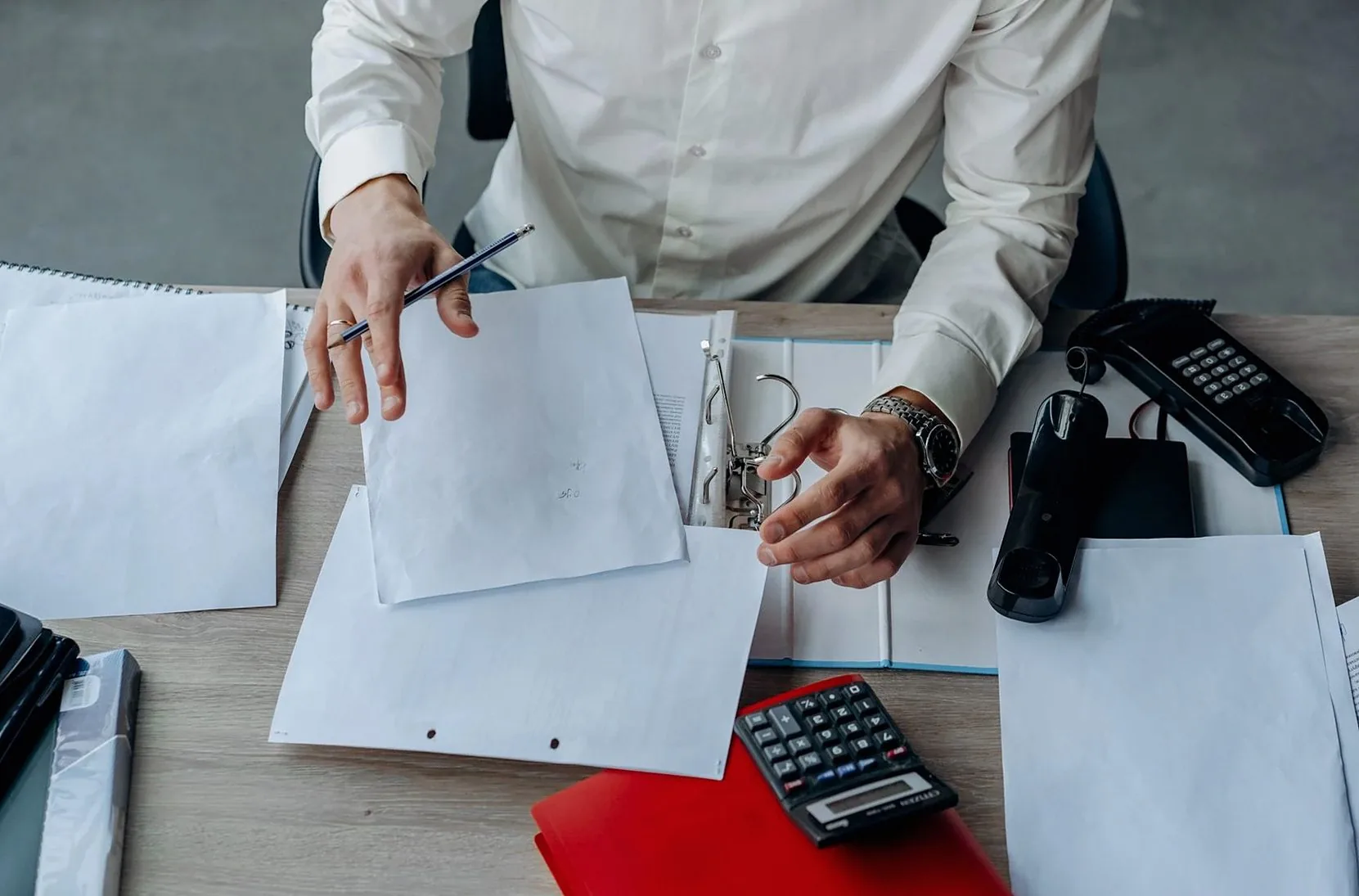 Person in a white shirt sorting paper documents at a desk, with a calculator and a red folder